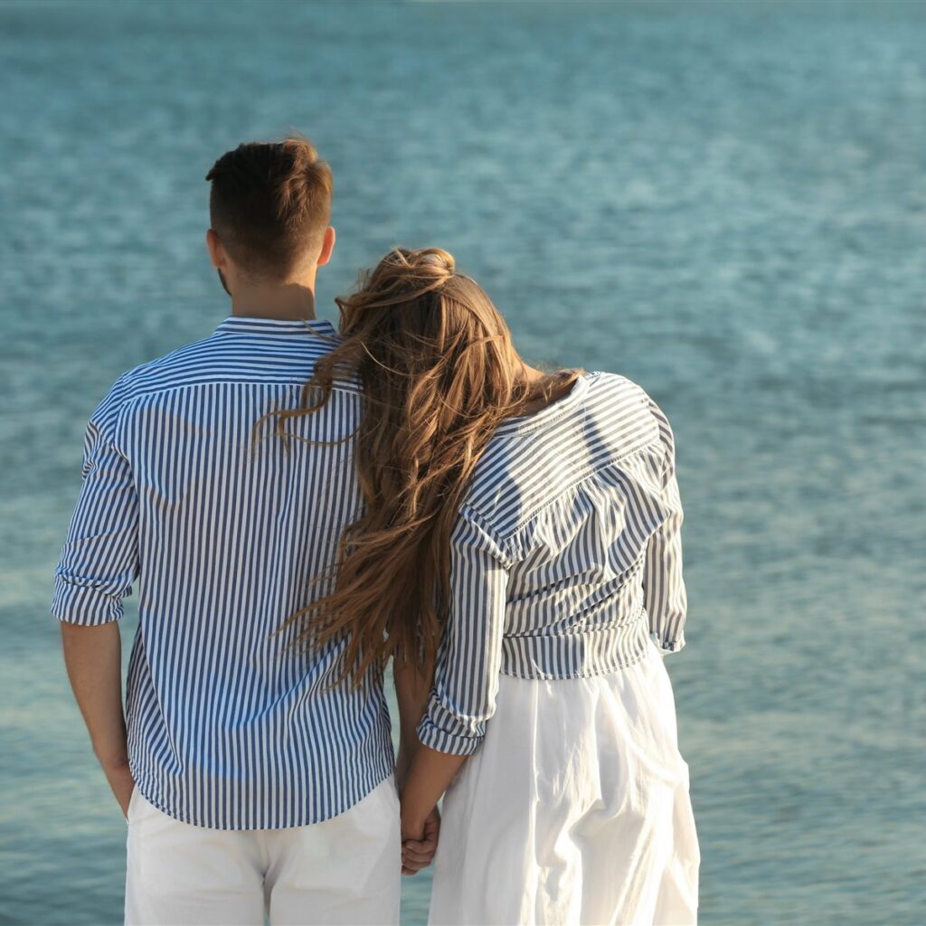 A couple in matching striped shirts seen from behind, holding hands and leaning on each other while looking at the sea